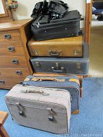 Photo showing five vintage luggage pieces stacked near a dresser. Variations in size, color, and material visible.