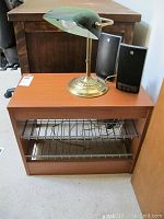 Wood shoe rack with brass and green glass banker lamp and two black speakers on top.