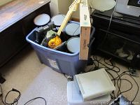 Overall view of bin containing multiple game controllers and guitar controller. Xbox 360 console visible on floor beside bin.