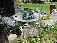 Wide shot showing round white wrought iron patio table with rust spots on top and four matching patio chairs arranged around it in an outdoor garden setting.