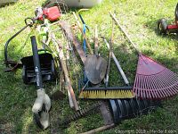 Photo showing assortment of yard tools laid on grass including rakes, shovels, broom, leaf blower, weed wacker, tomato cages, and snow shovel.