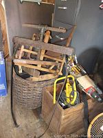 Lot with combination of wooden hand tools including bow saws stacked in basket, small wooden box holding yellow power drill and cord in a storage area