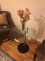 Front view of the glass top table with a square transparent surface and a vase with flowers placed on top. Surrounding furniture visible.