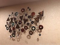 Side angle view of the metal wall sculpture hanging on a beige textured wall, showing the sculpture's dimensionality and mix of colors and shapes.