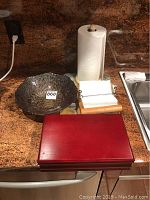 Full lot on granite countertop including a red wooden tea box, black decorative bowl, wooden napkin holder with napkins, and small cutting board next to a paper towel roll and sink.