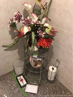 Full view of chrome bathroom table with two glass shelves, artificial flowers in white vase, small garbage can, soap dish, toilet paper holder, and napkin holder all grouped in bathroom corner.