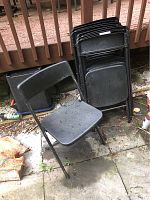 Group of folded and one open black metal folding chairs with plastic seats and backs outside on concrete patio next to wooden deck.