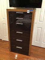 Front view of tall dresser showing six dark brown glossy drawers with metal handles, wood-grain walnut finish on sides and top.