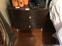 Front view of one night table showing curved front, two drawers, wood grain and metal feet