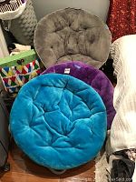 Three plush folding floor chairs stacked or leaned together showing teal, purple, and gray colors and tufted cushioned seats.