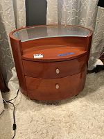 Front view of modern nightstand showing curved wooden body, two drawers with silver knob handles, and clear glass tabletop.