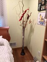 Full view of tall dark glass vase filled with decorative branches and reeds, standing beside bed and wall vents