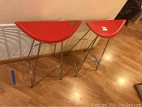 Two modern bar stools with crescent-shaped red leather seats and chrome metal frames shown from the side and front, placed on wooden floor against a wall.
