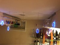 Photo of two metal wall mounted wine glass racks with multiple hanging glasses in different colors, blue circular LED lights hanging from ceiling, and shelves with liquor bottles underneath.