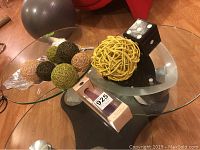 Photo shows a clear glass triangular dish holding assorted rattan decorative balls in natural colors and a yellow ball, a black square holder with glass coasters, and an aluminum leaf-shaped dish, all arranged on a wooden floor.