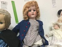 Front view of porcelain doll seated, showing detailed face, curly hair, and blue coat with red buttons.