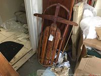 Vintage wooden sled with metal runners leaning against a wall indoors, showing front and side angles.