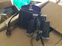 Vintage AMC binoculars with black textured body, silver knob adjustments, and leather strap beside matching black leather carrying case with AMC logo visible inside lid.