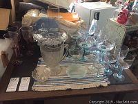 Varied assortment of clear stemware with colored stems, large cut glass pitcher and cake plate arranged on table.