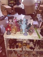 Photo shows 14 assorted glass vinegar cruets in various colors and sizes on white wooden surface. Background includes boxes and household items.