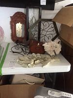 Table display with assorted wall art items including mirrors, metal framed glass panel, wooden box, coral sculpture, resin angel, and metal decorative pieces.