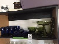 Shelf with 9 blue glasses and 7 stacked blue plates on left, and 5 olive green glass pieces including bowls, a large pedestal dish, and starfish shaped dishes on right.