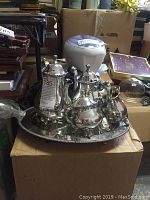 Photo showing the entire five-piece silver-plated tea and coffee set arranged on a round serving tray, placed on a cardboard box among other items in storage.