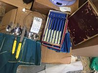 A collection of stainless steel flatware with yellow-handled knives wrapped in a cloth roll alongside boxed steak knives and a glass plate
