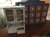 Green wall mount cabinet and small white wall mount cabinet shown together on a surface, the green one having 12 drawers with white knobs, the white one having mesh doors and small drawers below.