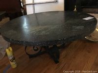 Side view showing round granite tabletop and decorative metal base on wood floor with a yellow Pringles can for scale.
