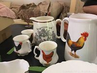 Photo showing two pitchers and three mugs on a table, featuring rooster and chicken designs.