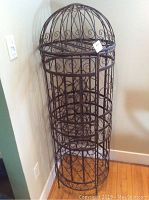 Full view of tall cylindrical metal wire wine rack with dome top resembling a bird cage, standing on hardwood floor next to wall.