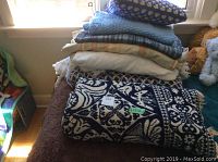 Stack of folded bedding items including a dark blue and white patterned chenille bedspread and assorted sheets and afghans on a brown surface.