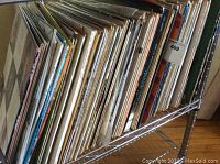 Photo of a large collection of vinyl LP records stacked side by side on a metal shelf, covers partially visible showing a variety of artists and titles.