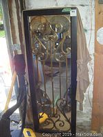 Full view of rectangular iron mirror gate panel with black frame and gold iron scrollwork detail, leaning against a wall.