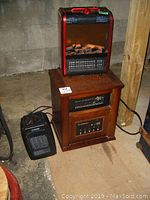 All three heaters positioned on a floor in a basement: red Garrison heater on a wood-like heater base, small Likewise heater to the left close by. Bright lighting from camera flash.