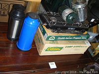 Photo showing two thermoses, camping stoves stacked on boxes, propane canister, and aluminum coffee pot