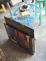 Photo showing the blue painted metal table with floral pattern, two framed artworks leaning against it, and a wooden ashtray on top.