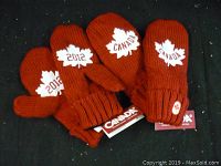 Two pairs of red knit mittens each displaying a white maple leaf patch with '2012' and 'Canada' text, arranged on a black surface.