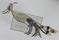 Full view of vintage TARA peacock brooch resting on a clear display stand showing the full length and metal details under multi colored rhinestones.
