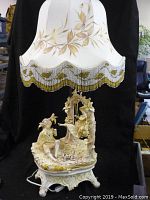 Full view of the lamp with detailed porcelain base and fabric shade with gold painted leaves and beaded fringe.