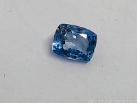 Close-up photo of the antique cut heated blue sapphire gemstone resting on a white background, showing its cushion shape, blue coloration, and faceting.
