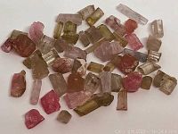 Photo showing a variety of rough tourmaline stones in multiple colors including pink, watermelon, blue, and greenish hues spread on a white surface.