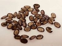 Top view of many oval calibrated colour change garnet stones scattered on a surface showing their various brown to purplish hues.