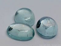 Three natural aquamarine cabochon cut stones arranged on flat surface, showing oval shapes and translucent light blue colors with some inclusions.