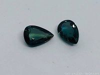 Close-up photo of two pear-shaped pear modified mixed cut blue/green natural sapphires on a plain background.