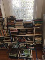 Three shelves filling the space by a window with an assortment of books mostly stacked vertically and some horizontally. Visible titles relate to travel, history, and religion.