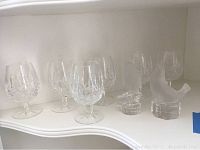 Front view of six Waterford Lismore crystal wine glasses and two Nachtmann crystal figurines on white shelf.