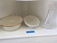 Three Lenox white china serving bowls and one Waterford crystal wine decanter with stopper on a white shelf.