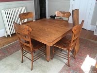 Full view of square golden oak dining table surrounded by four matching oak chairs with carved backrests, visible wear on wood surfaces